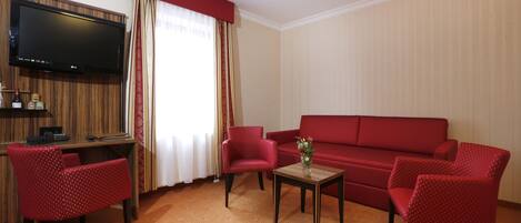 A hotel room with a TV, red furniture, a small table with a vase, and a window with curtains.