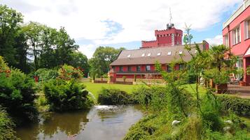 A castle-like building with a red exterior and a tower, surrounded by lush greenery and a small pond.
