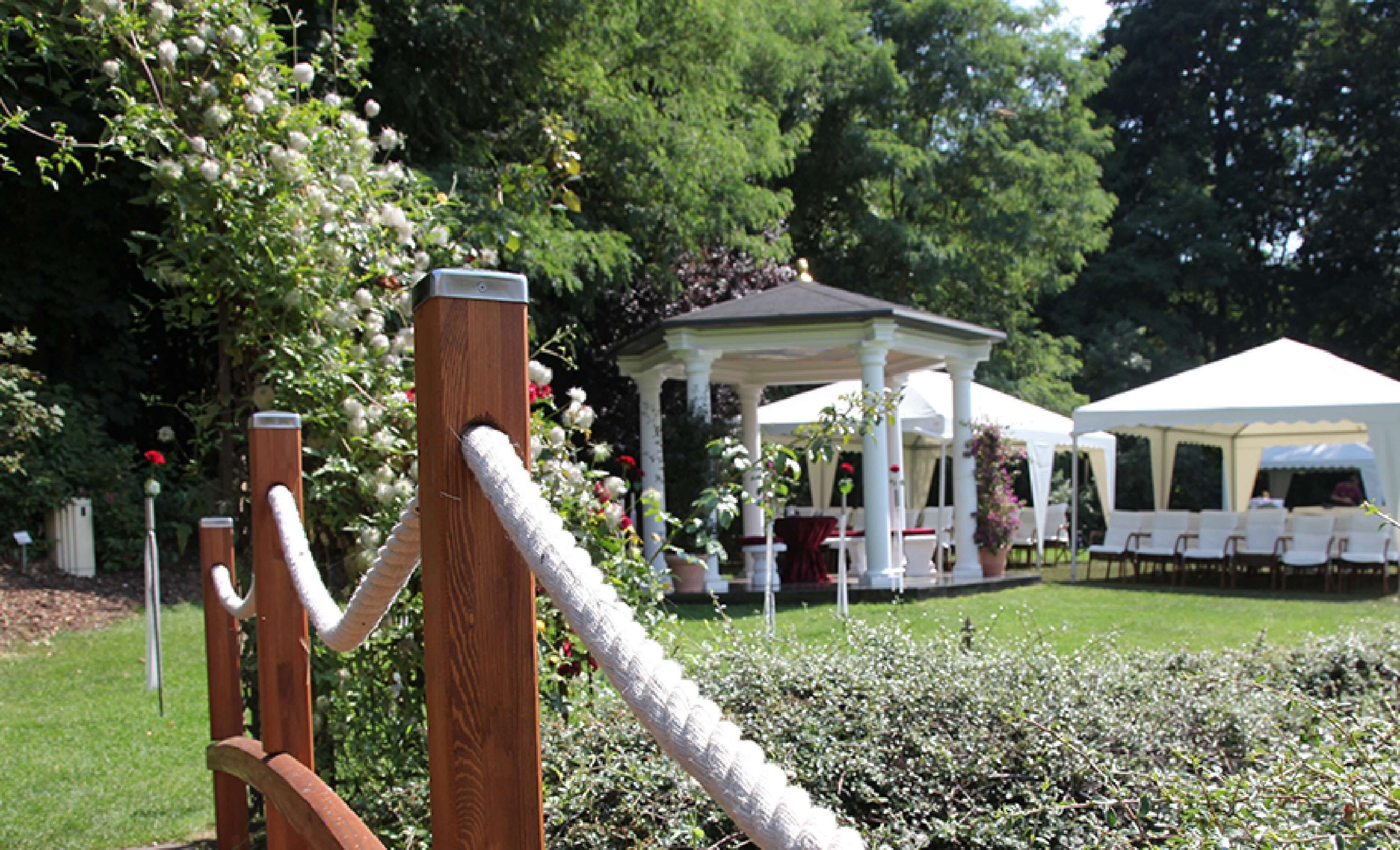 A gazebo with a white roof and columns, surrounded by lush greenery and flowering plants.