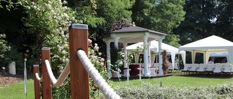 A gazebo with a white roof and columns, surrounded by lush greenery and flowering plants.
