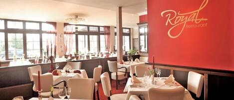 Interior de un restaurante con mesas dispuestas para comer, una pared roja con el nombre del restaurante y grandes ventanales que dejan entrar luz natural.