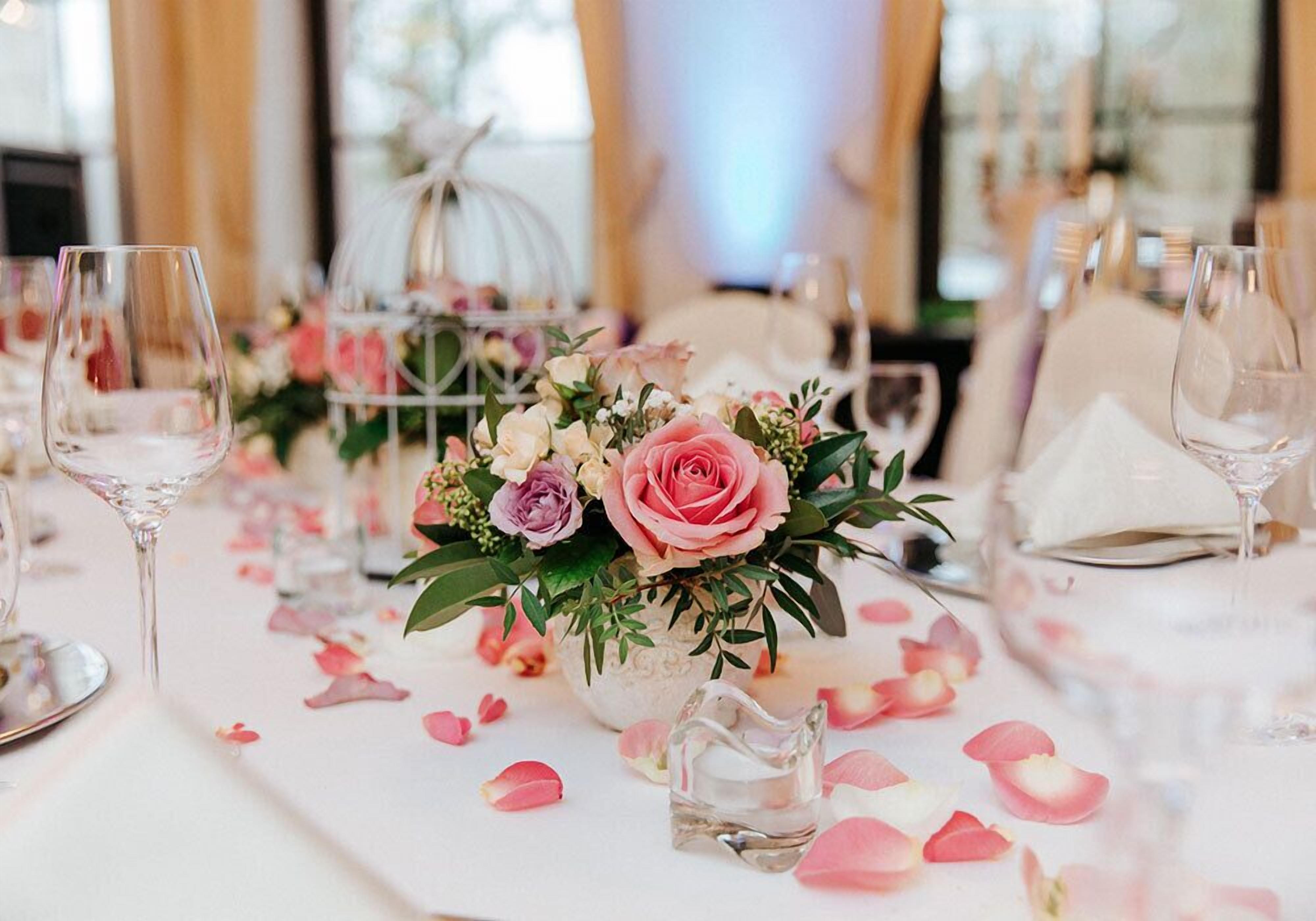 A table set with wine glasses, a centerpiece of pink roses, and scattered rose petals.