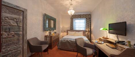 A hotel room with a bed, two chairs, a desk with a computer, a TV, and a chandelier.