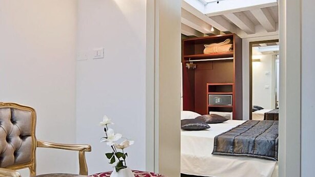 Quarto com cama, uma cadeira e uma mesinha com um vaso de flores.