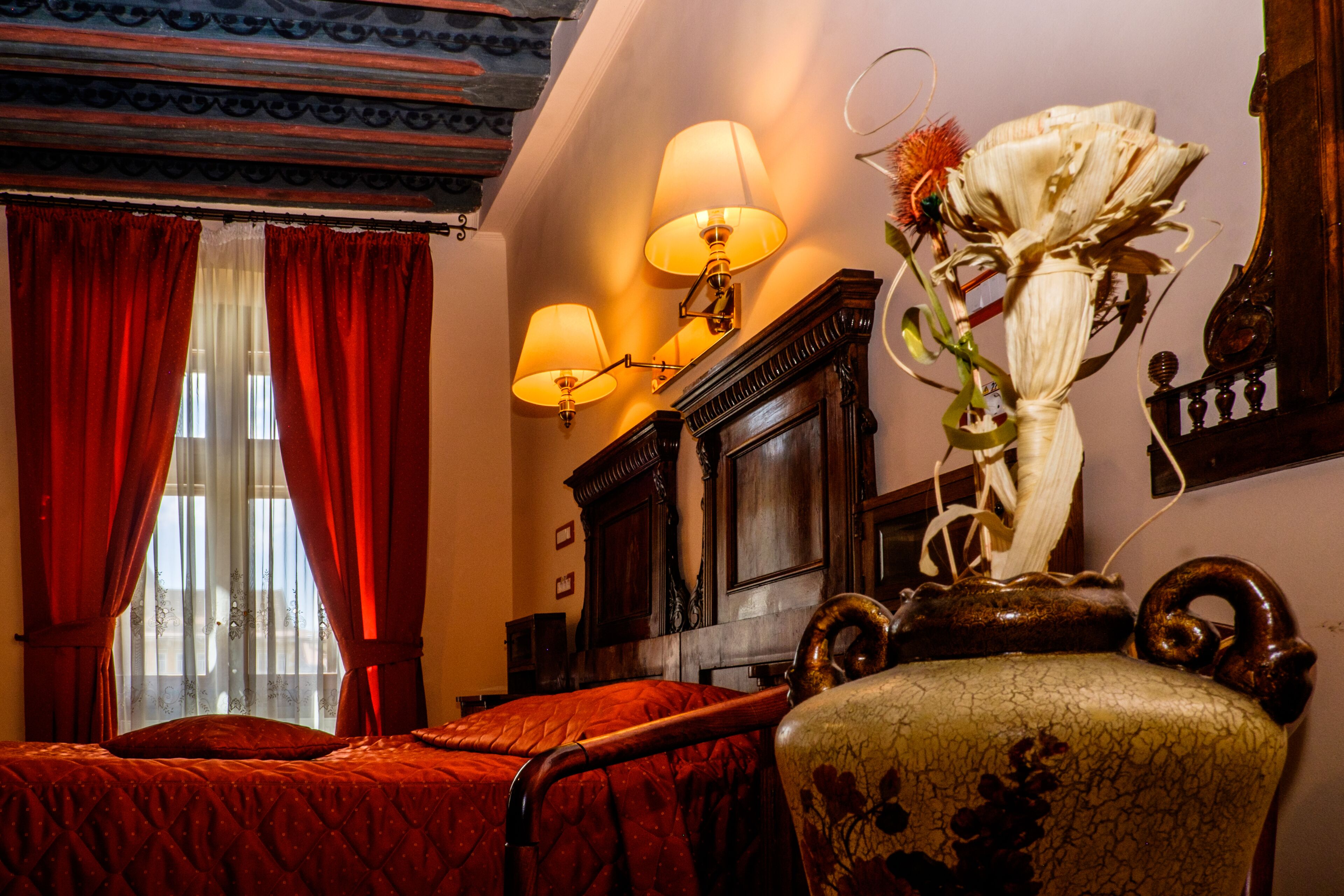Quarto com cama, cortinas vermelhas, um vaso de flores e luminárias na parede.