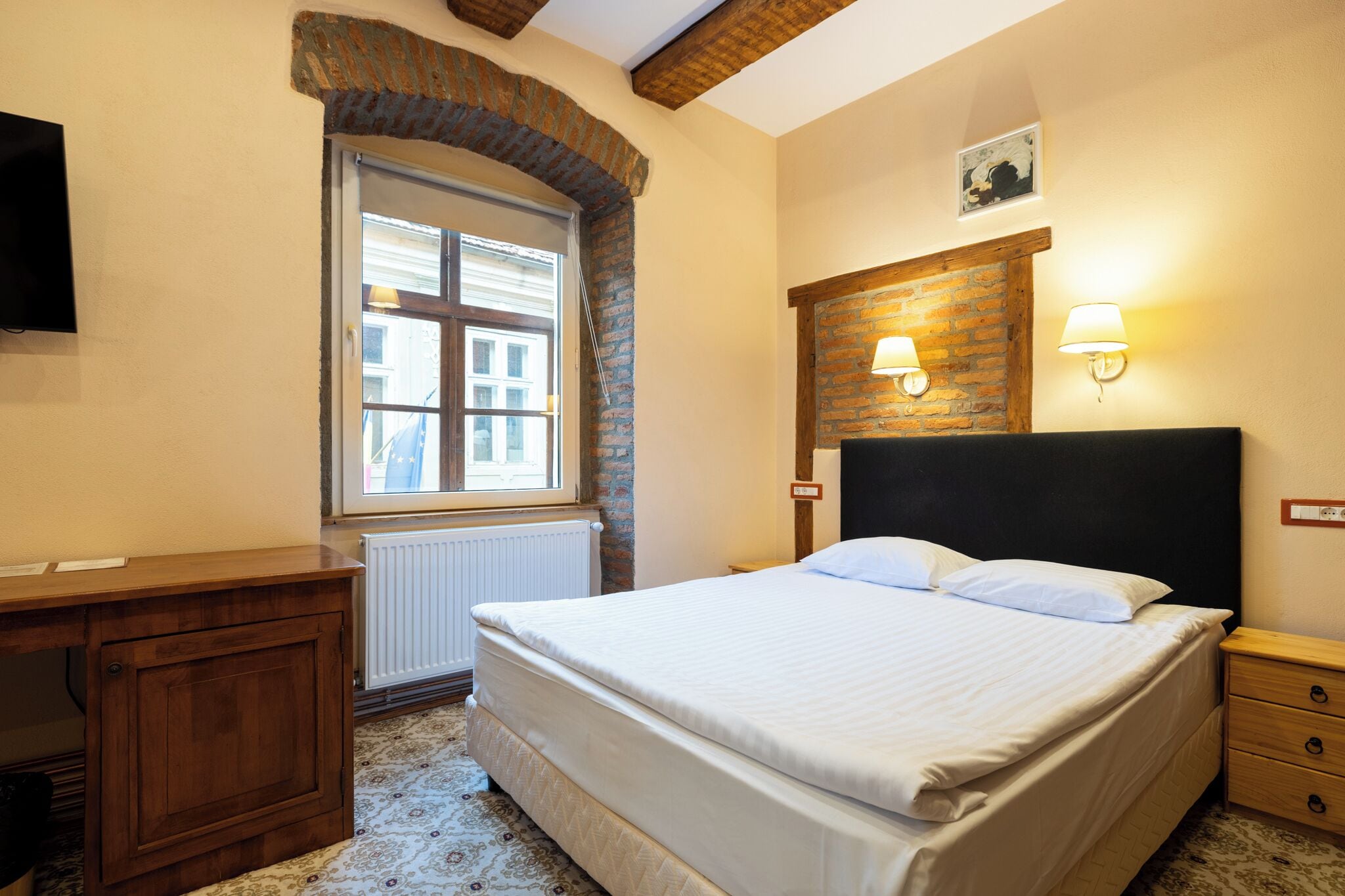A bedroom with a bed, wooden headboard, a window, a radiator, a desk, and a television.