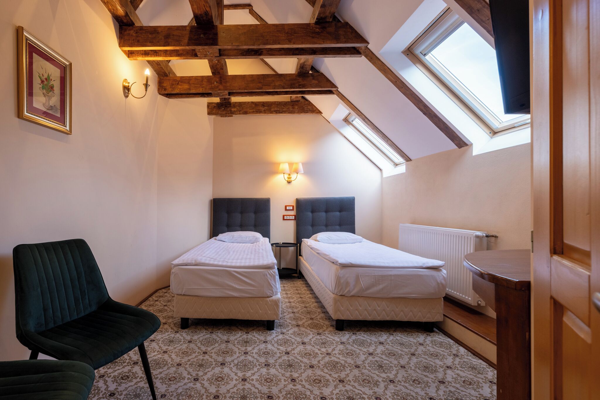 Una habitación pequeña y acogedora con dos camas individuales, una silla, una lámpara de pared y un tragaluz.