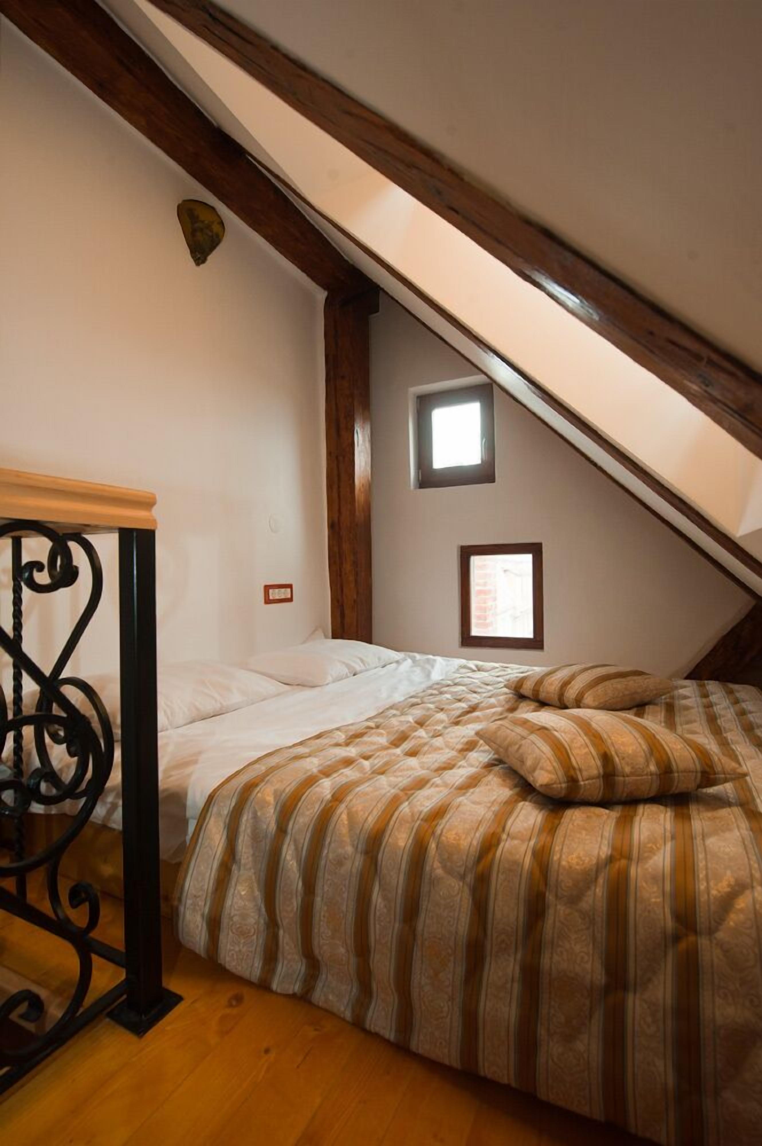 Quarto com teto inclinado, cama com roupa de cama listrada, uma mesinha e duas janelas.