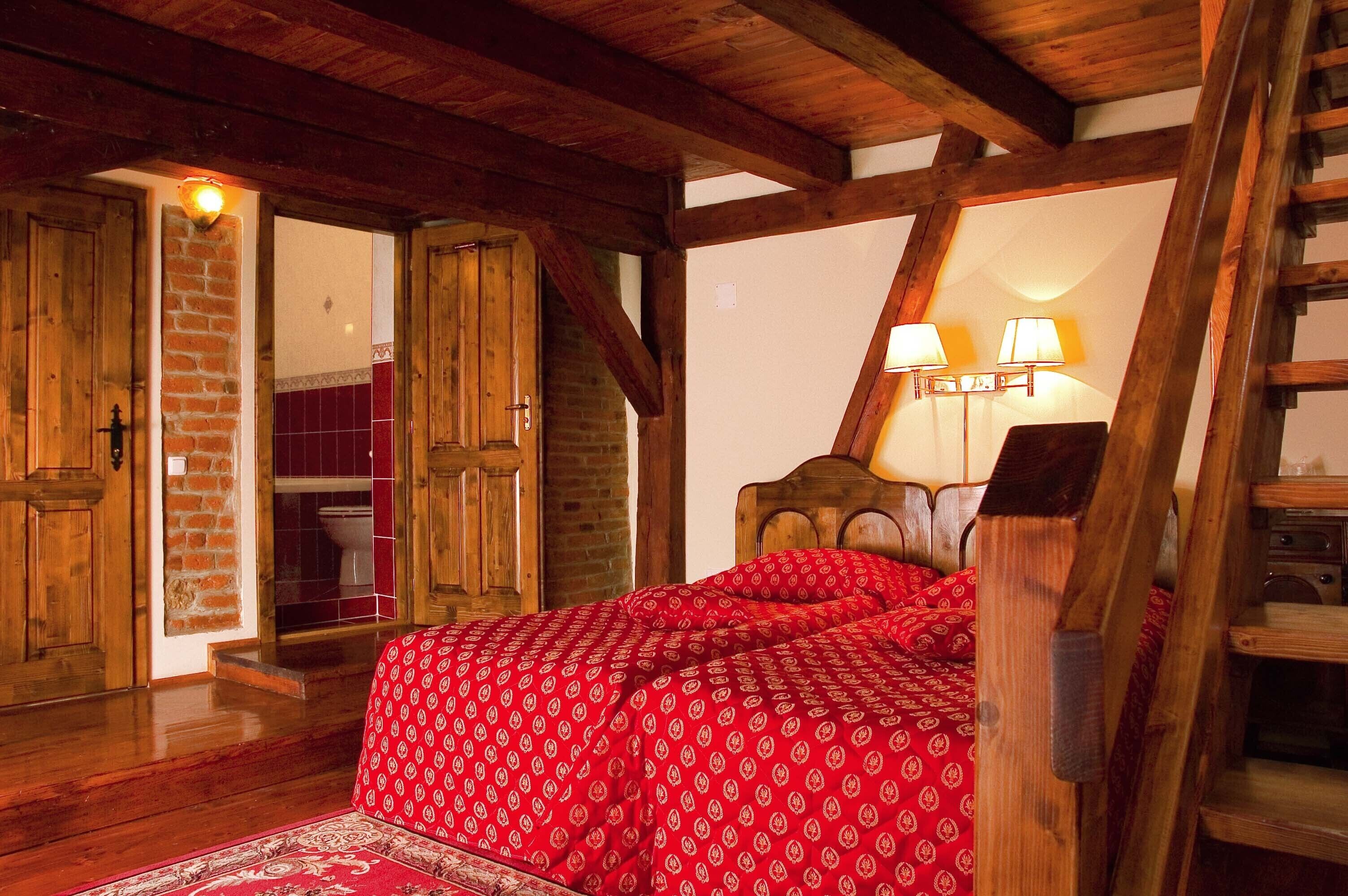 A bedroom with a wooden ceiling, a bed with red bedding, a brick wall, and a wooden ladder.