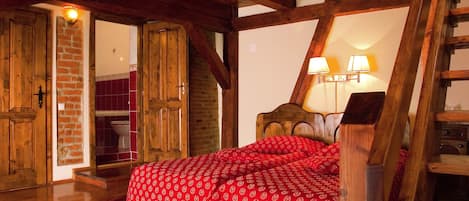 A bedroom with a wooden ceiling, a bed with red bedding, a brick wall, and a wooden ladder.