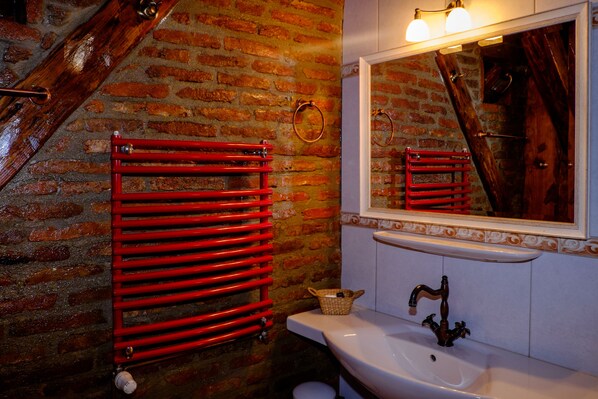 Un baño con pared de ladrillo, toallero rojo, lavamanos blanco y espejo con reflejo de una puerta de madera.