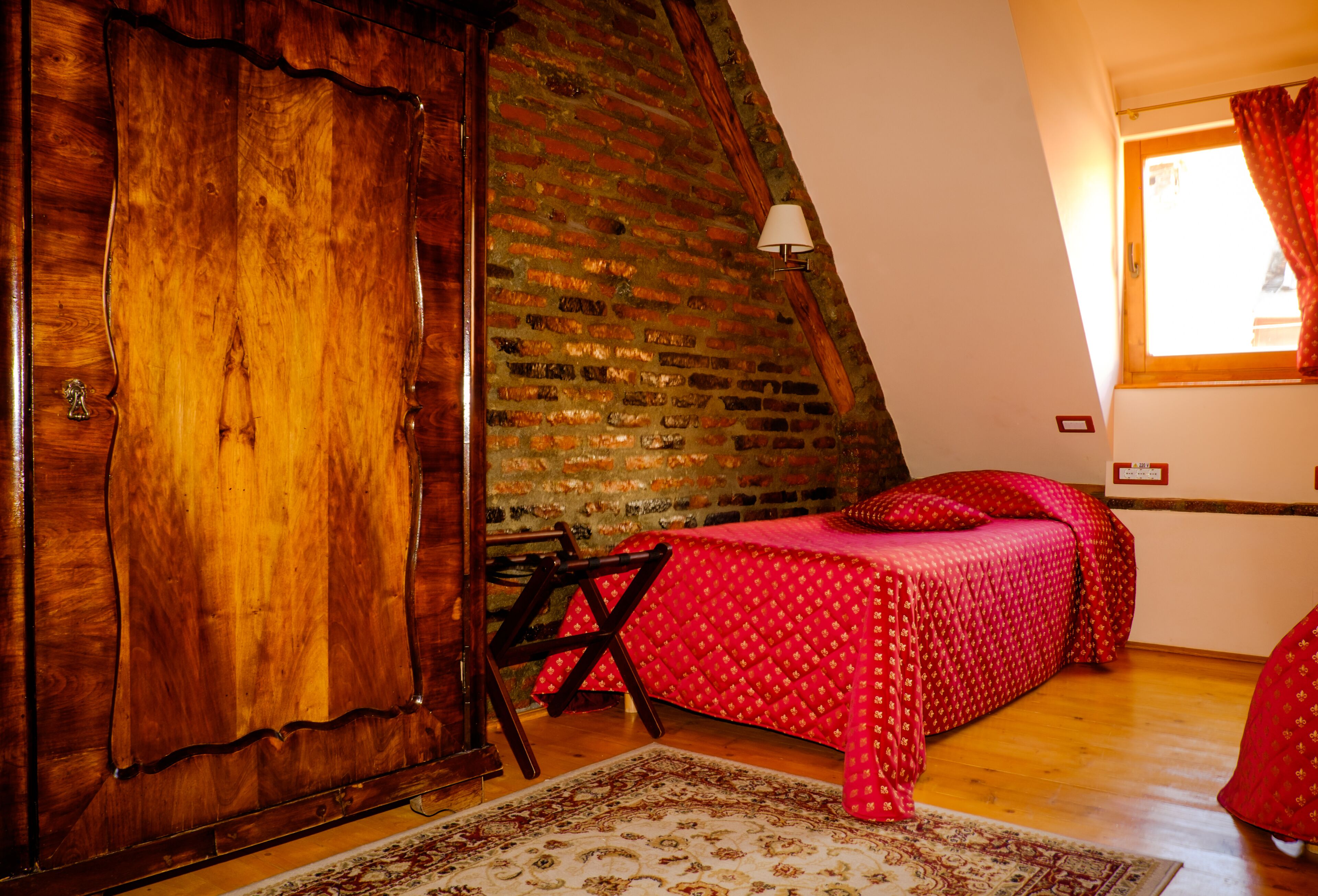 Une chambre avec une armoire en bois, un lit recouvert d’une courtepointe rouge, une fenêtre avec des rideaux et un mur en briques.