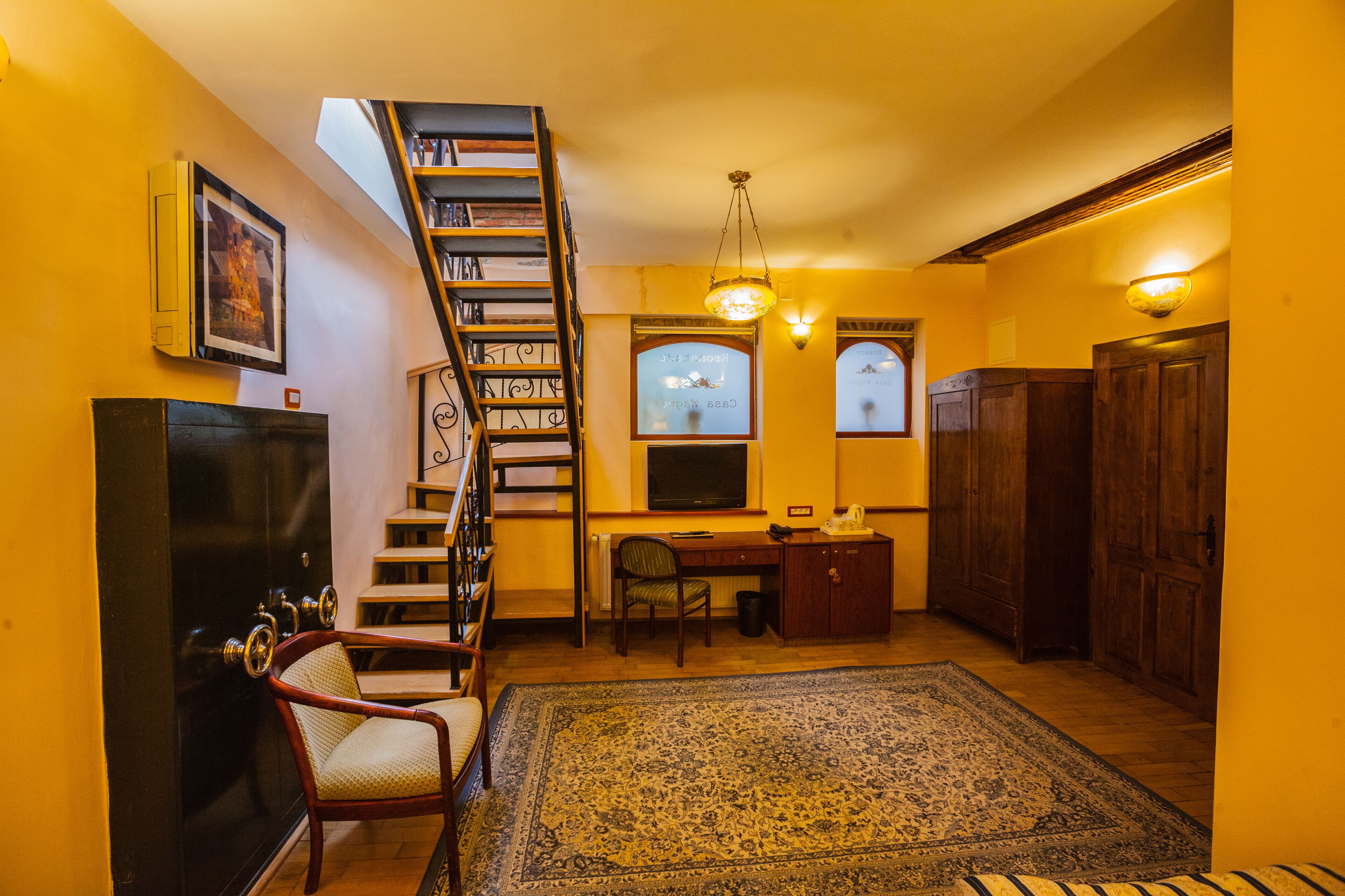 A room with a wooden staircase, a desk with a chair, a radiator, a large rug, and a television.