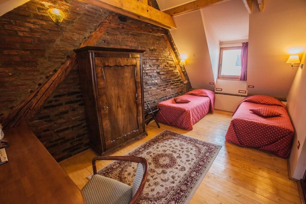 Un dormitorio acogedor en el ático con vigas de madera a la vista, un armario rústico, dos camas individuales con ropa de cama roja, una silla de madera y una alfombra estampada.