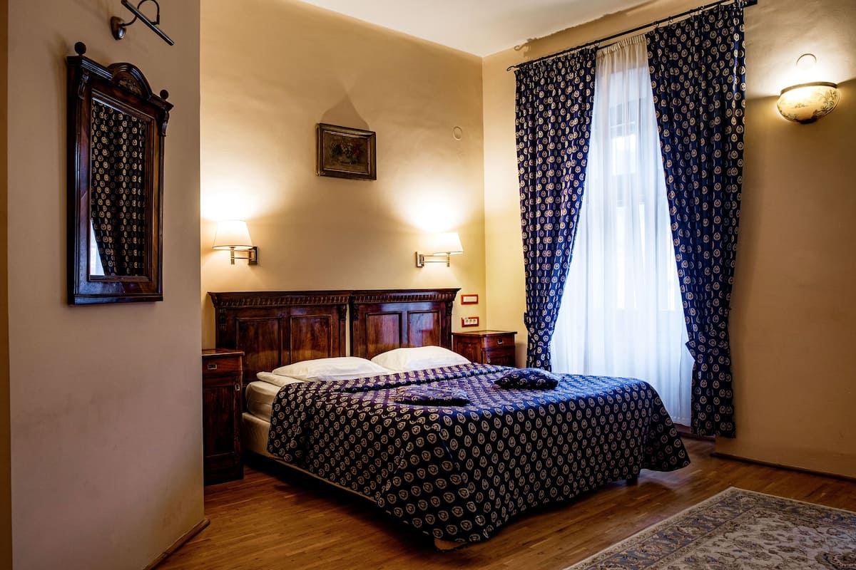 A bedroom with a wooden bed, two bedside tables, a nightstand, a wall-mounted lamp, a mirror, and a patterned bedspread.
