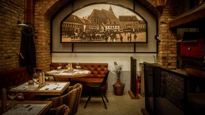 A restaurant interior with brick walls, a large arched window displaying a historical street scene, wooden tables and chairs, and a fireplace. - Casa Wagner Brasov (Brasov)