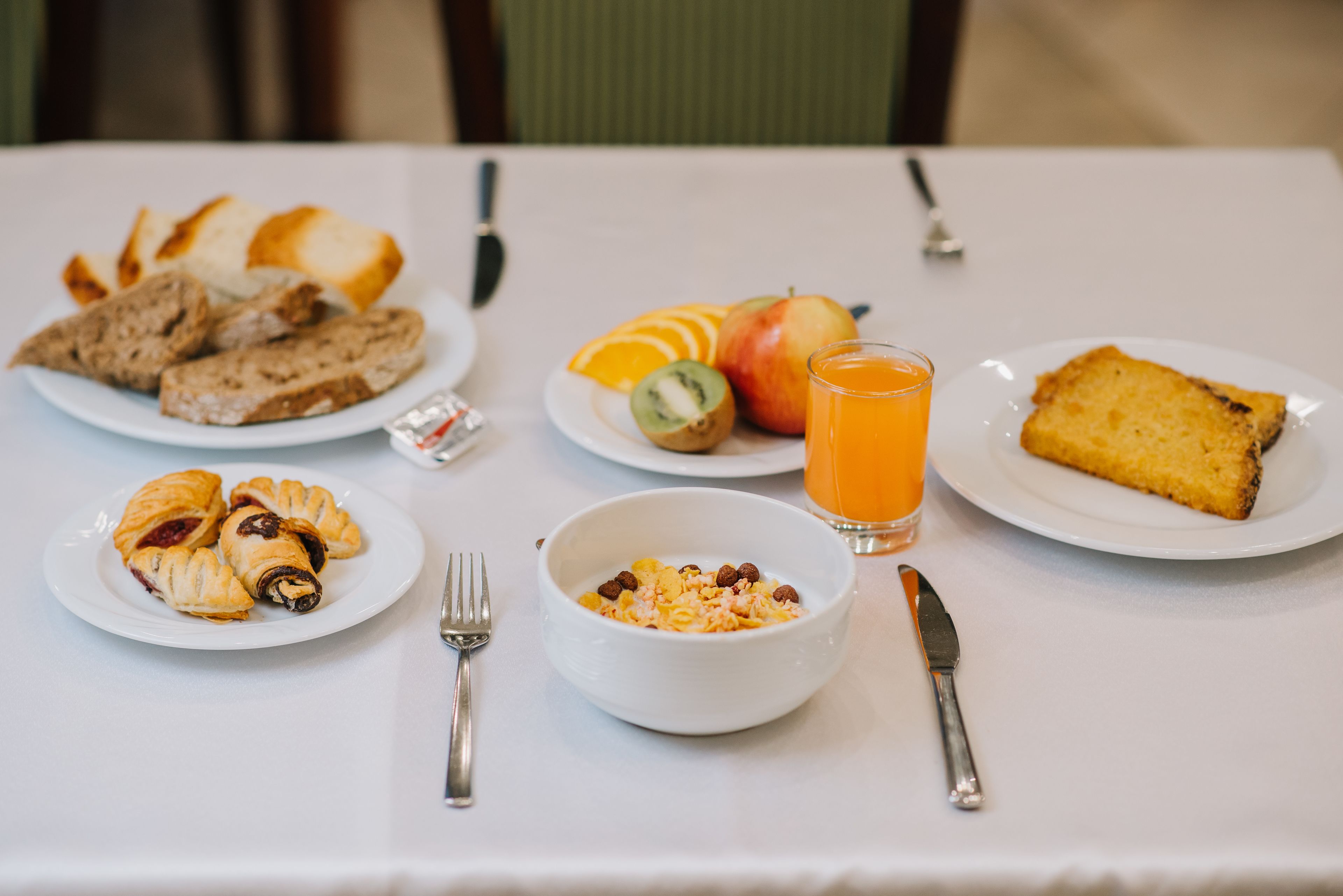 Une table de petit-déjeuner avec des assiettes de viennoiseries, des fruits, un bol de céréales et un verre de jus d’orange.