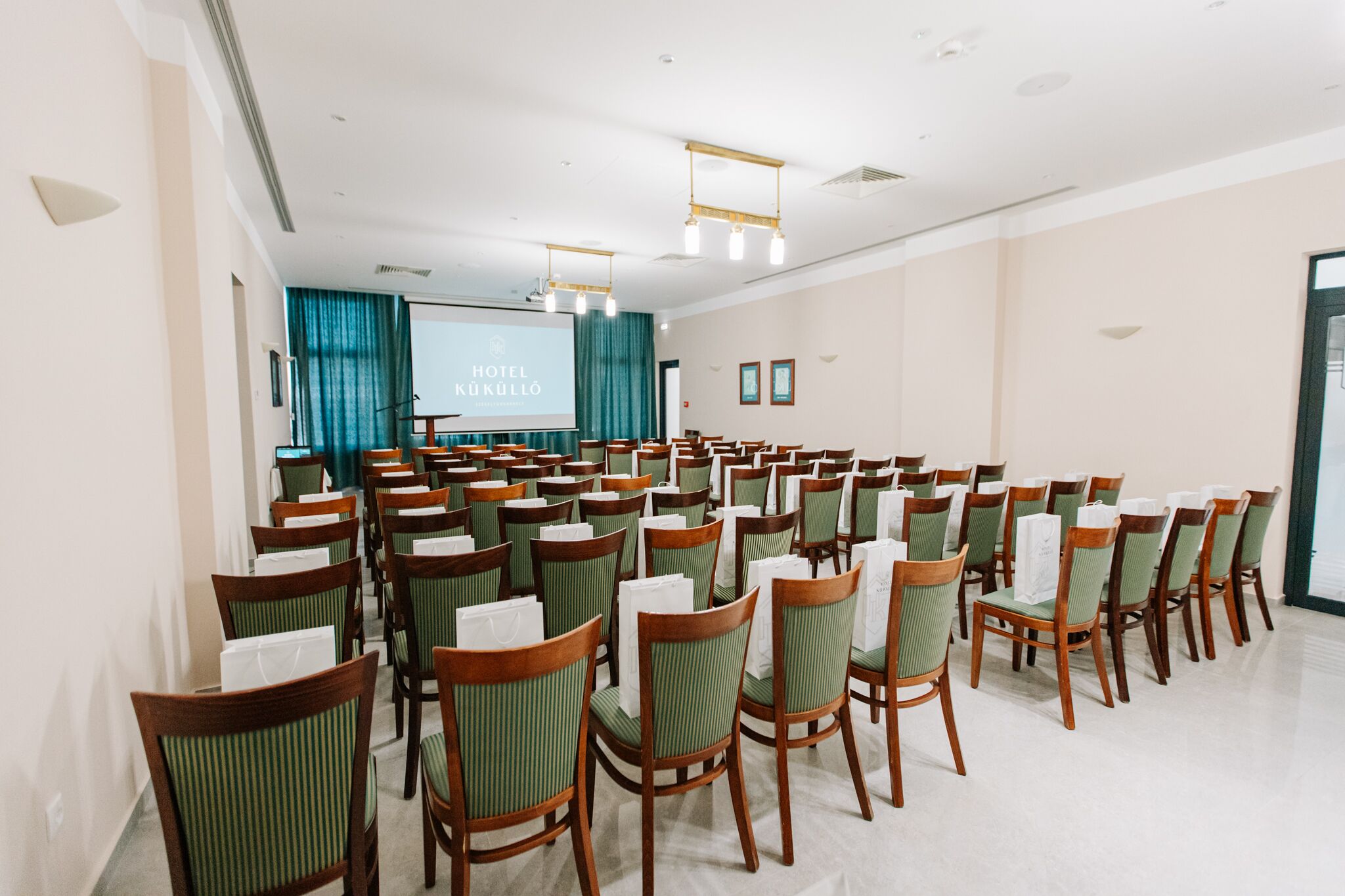 Un salón de conferencias con filas de sillas de madera y asientos tapizados en verde, una pantalla grande al frente y un proyector.