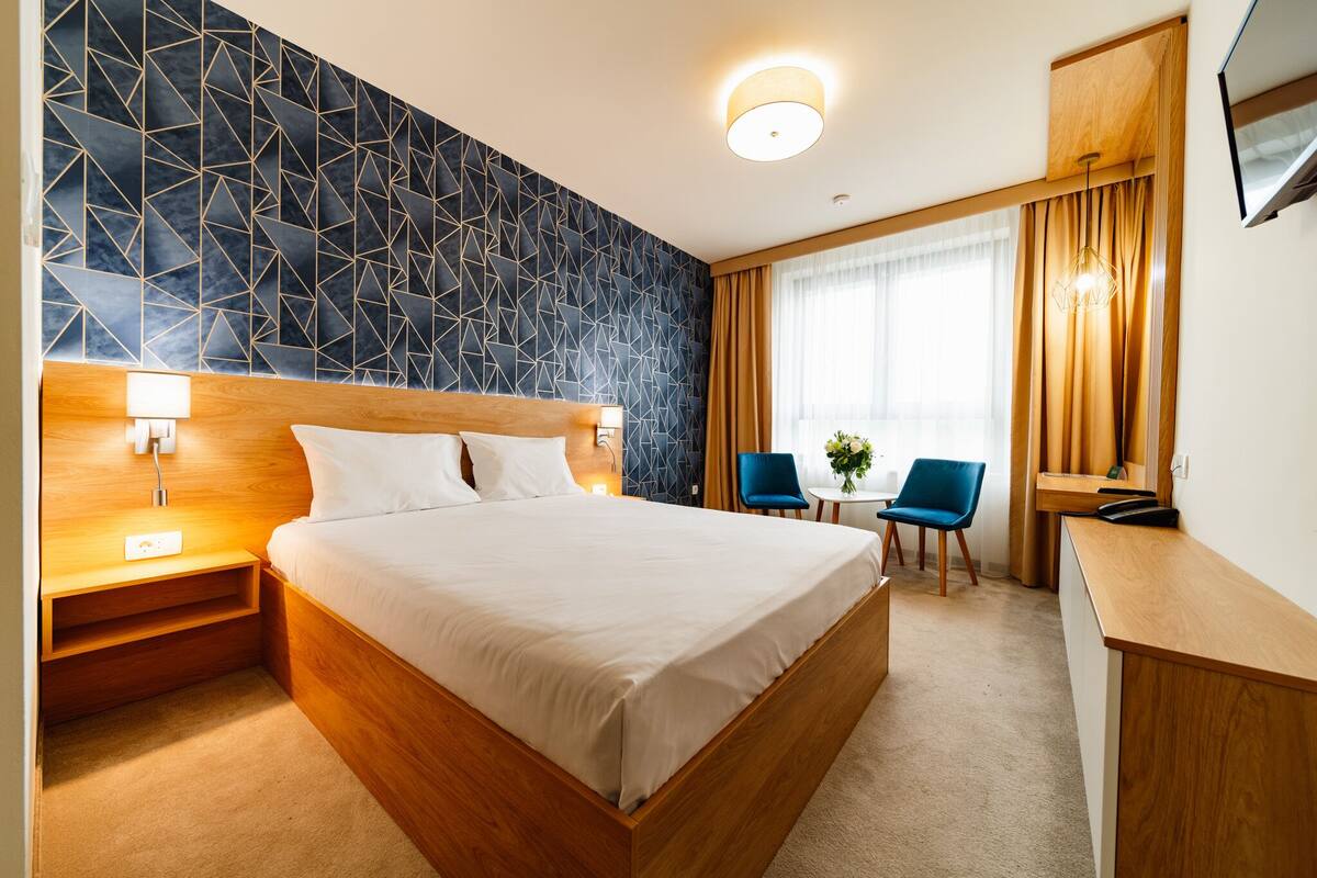 Ein modernes Hotelzimmer mit Wand in geometrischem Muster, einem hölzernen Bett, Nachttischen, einem Schreibtisch und einem Sessel.