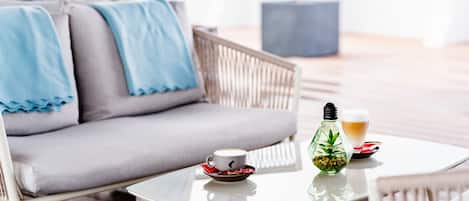 A modern outdoor seating area with a white table, a grey sofa with blue cushions, a glass coffee cup, a glass of beer, and a decorative bottle.