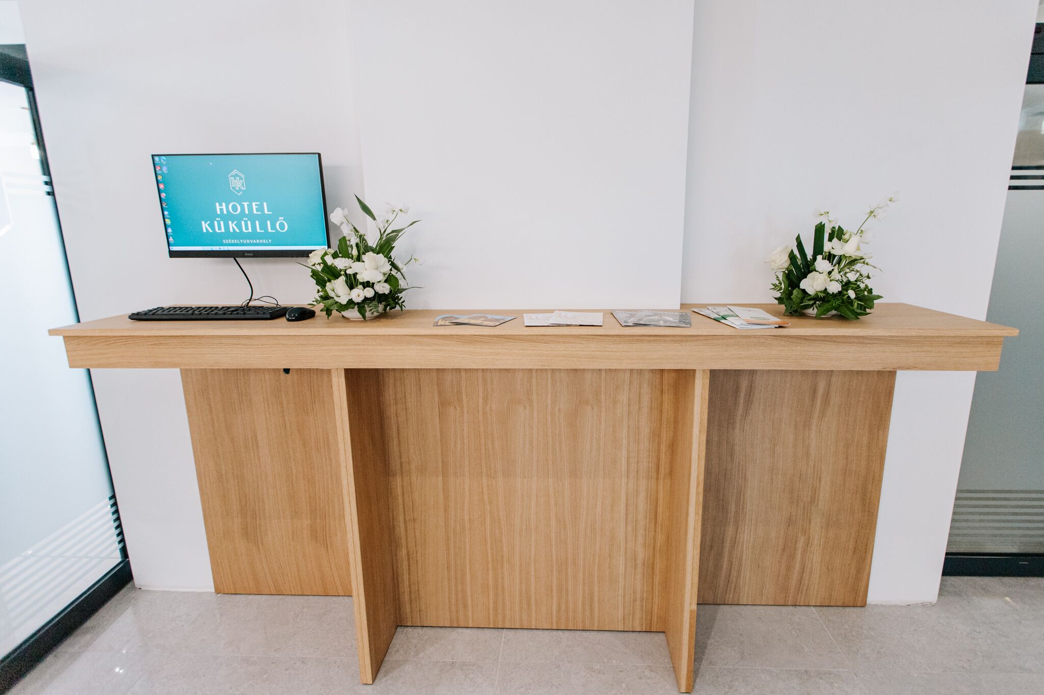 Ein Hotelrezeptionstresen mit einem Computermonitor, der 'HOTEL KUKÜLLO' anzeigt, einer Tastatur und zwei Blumenarrangements.