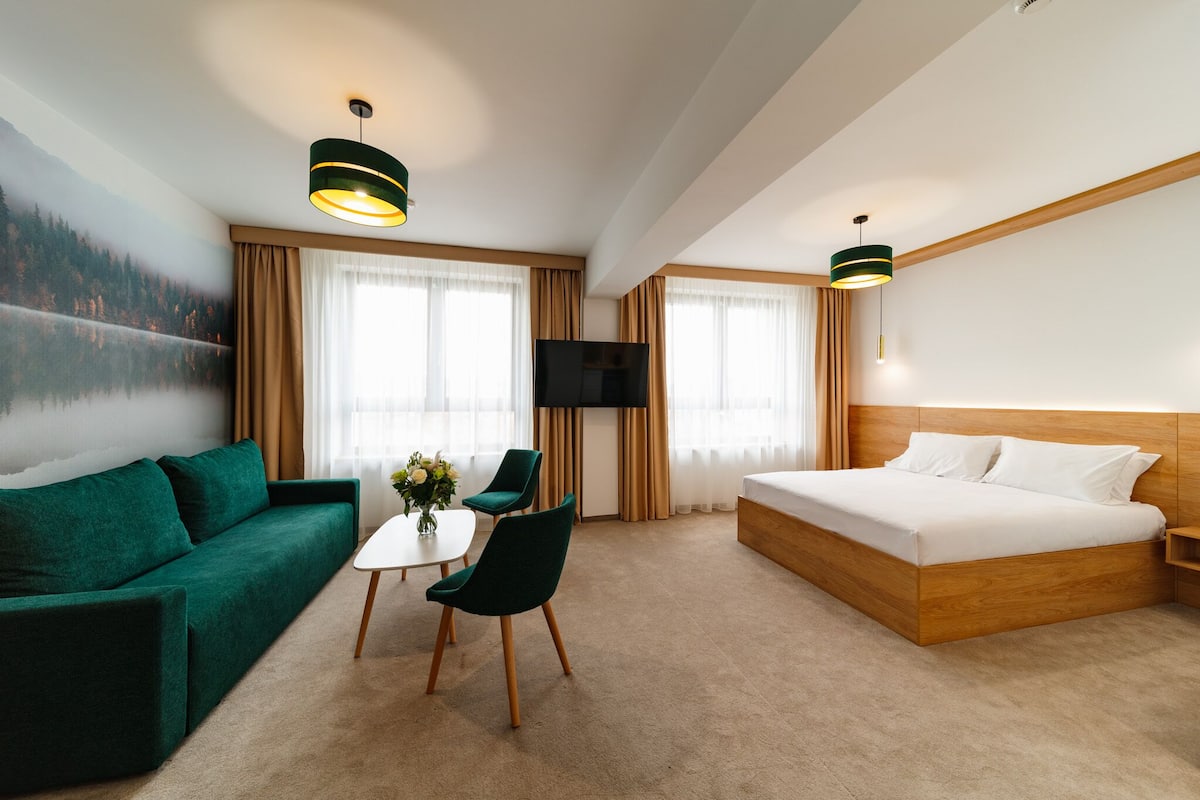 Ein modernes Hotelzimmer mit einem grünen Sofa, einem hölzernen Bett, einem Fernseher und einem Waldgemälde an der Wand.