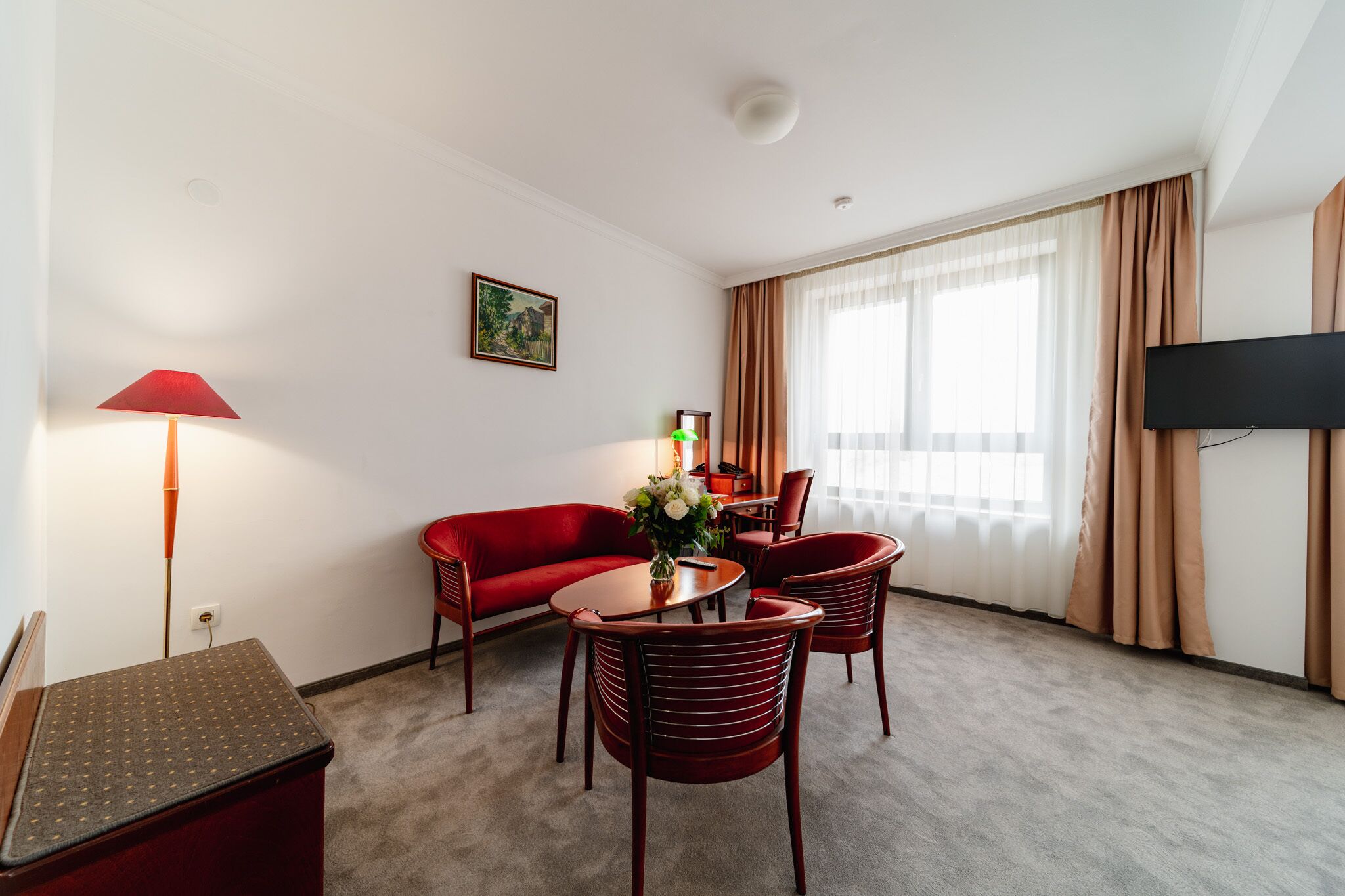 Una habitación de hotel con un sofá rojo, una mesa de comedor con sillas, un televisor montado en la pared y un ventanal con cortinas.