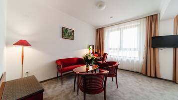 Una habitación de hotel con un sofá rojo, una mesa de comedor con sillas, un televisor montado en la pared y un ventanal con cortinas.