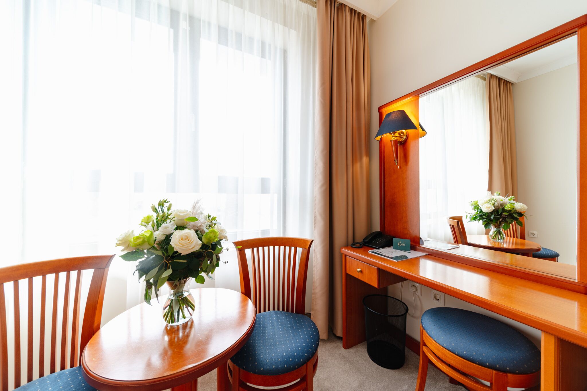 Habitación de hotel con escritorio de madera, dos sillas, un florero con flores, una lámpara y un espejo grande.