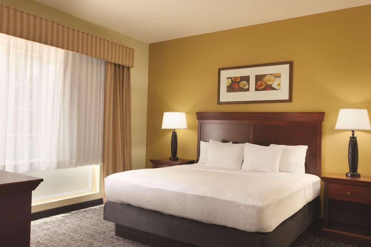 A hotel room with a large bed, two bedside tables with lamps, a window with curtains, and a framed picture on the wall.