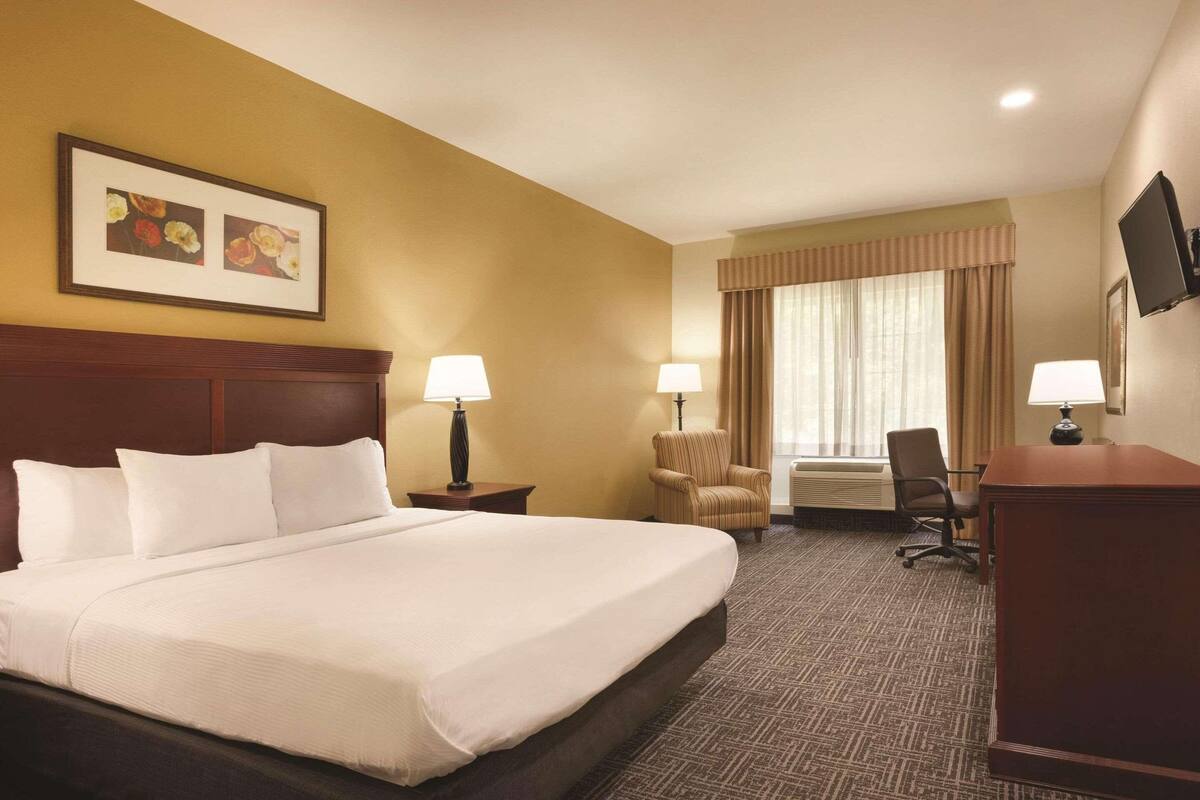 A hotel room with a large bed, two bedside tables with lamps, a desk with a chair, a television, and a window with curtains.