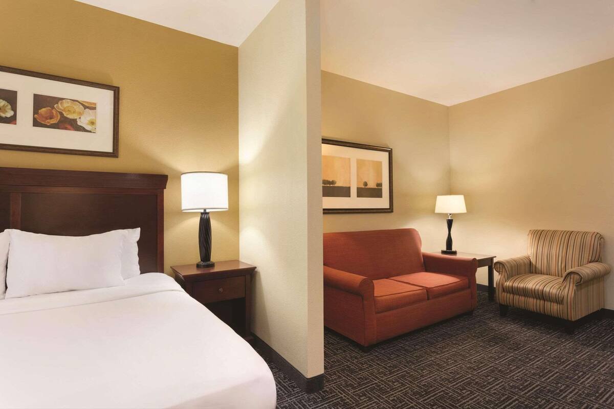 A hotel room with a bed, a nightstand with a lamp, a sofa, a striped armchair, and a framed picture on the wall.