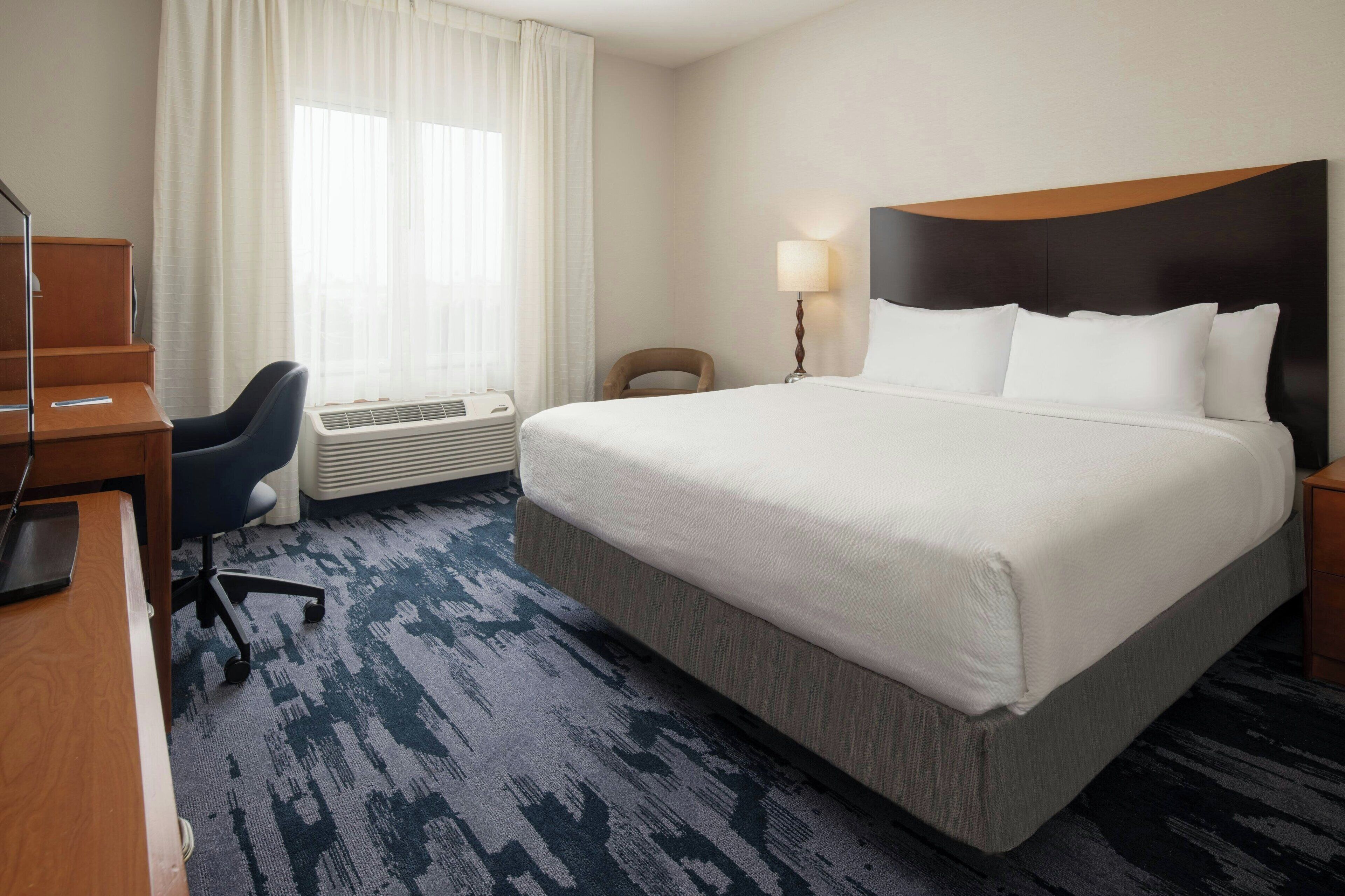 A hotel room with a large bed, a desk, a chair, a lamp, and a window with curtains.