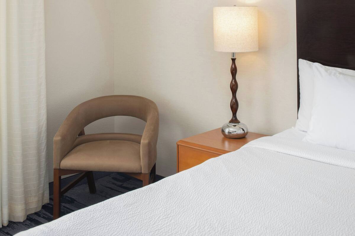 A hotel room with a bed, a chair, a nightstand with a lamp, and a curtain.