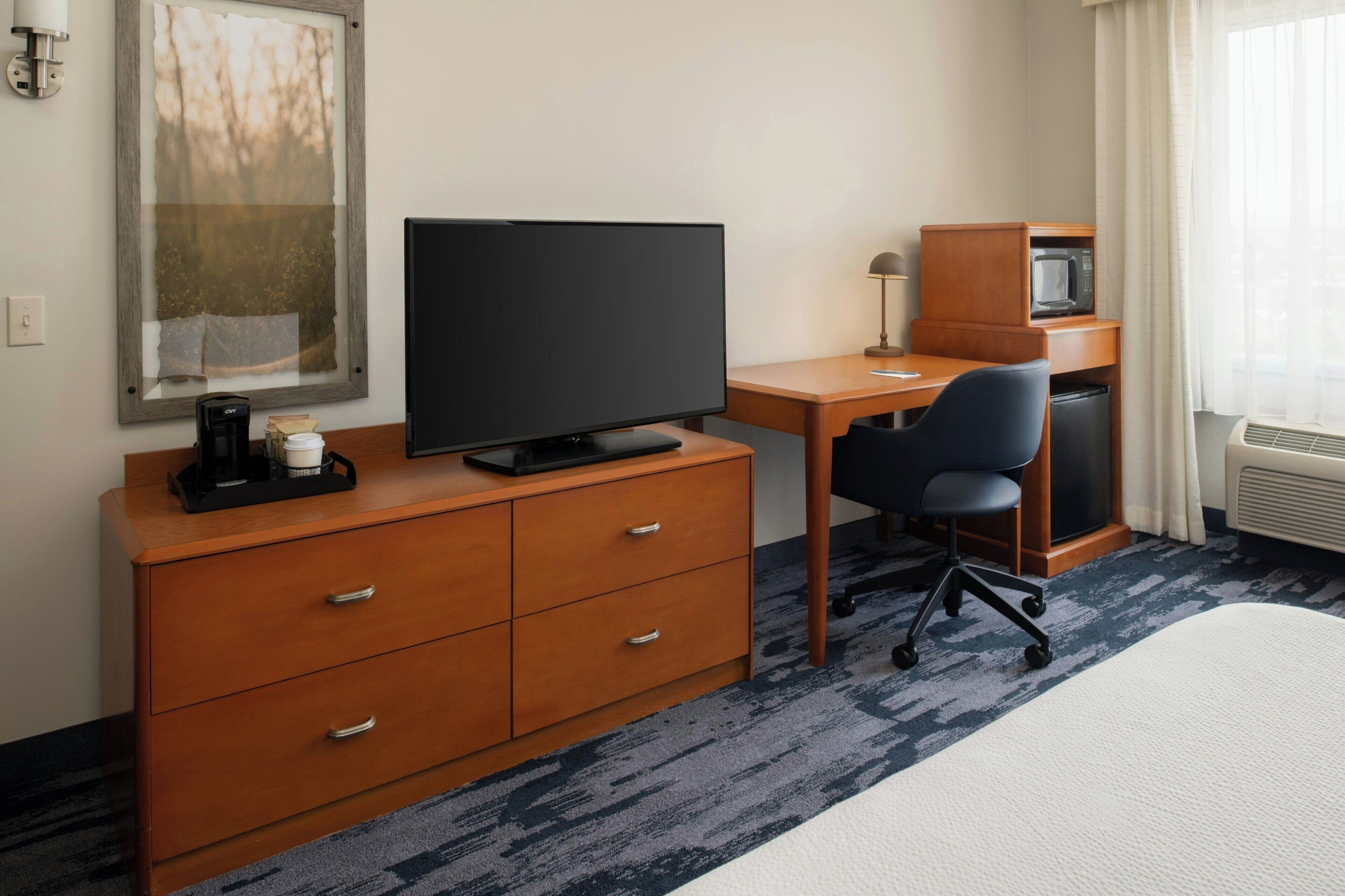 Quarto de hotel com escrivaninha, televisão, micro-ondas, cafeteira e uma cadeira.