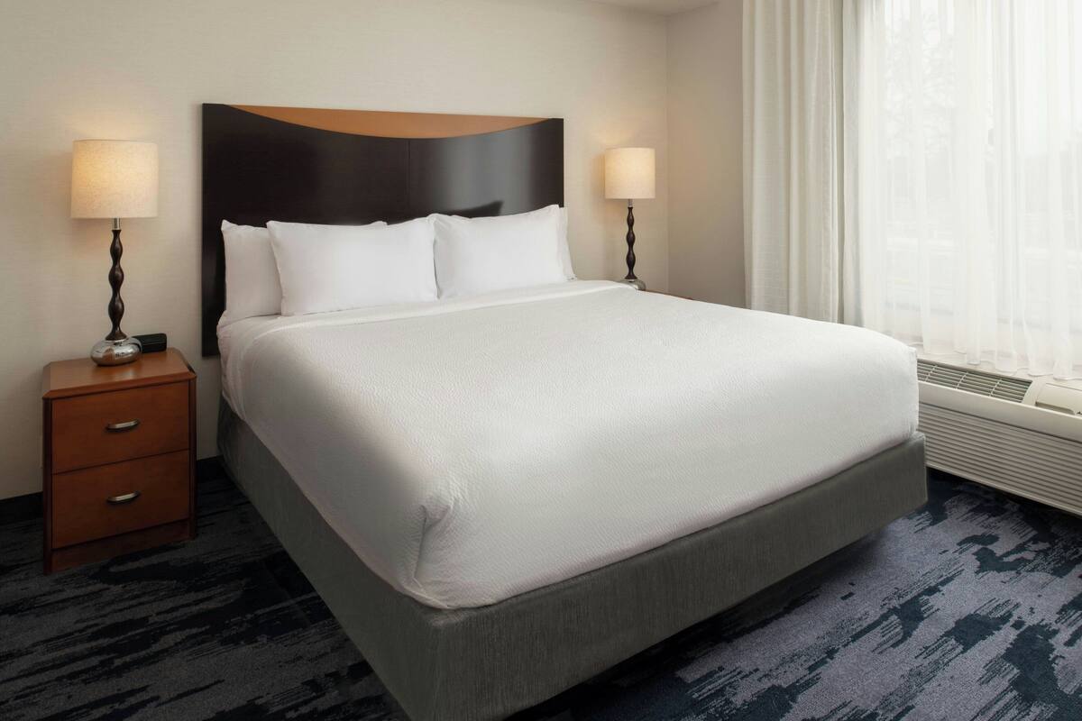 A hotel room with a large bed, two bedside tables with lamps, a dark headboard, and a view of the outdoors through sheer curtains.