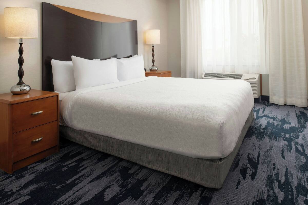 A hotel room with a large bed, two bedside tables with lamps, a wooden headboard, a window with curtains, and a carpeted floor.