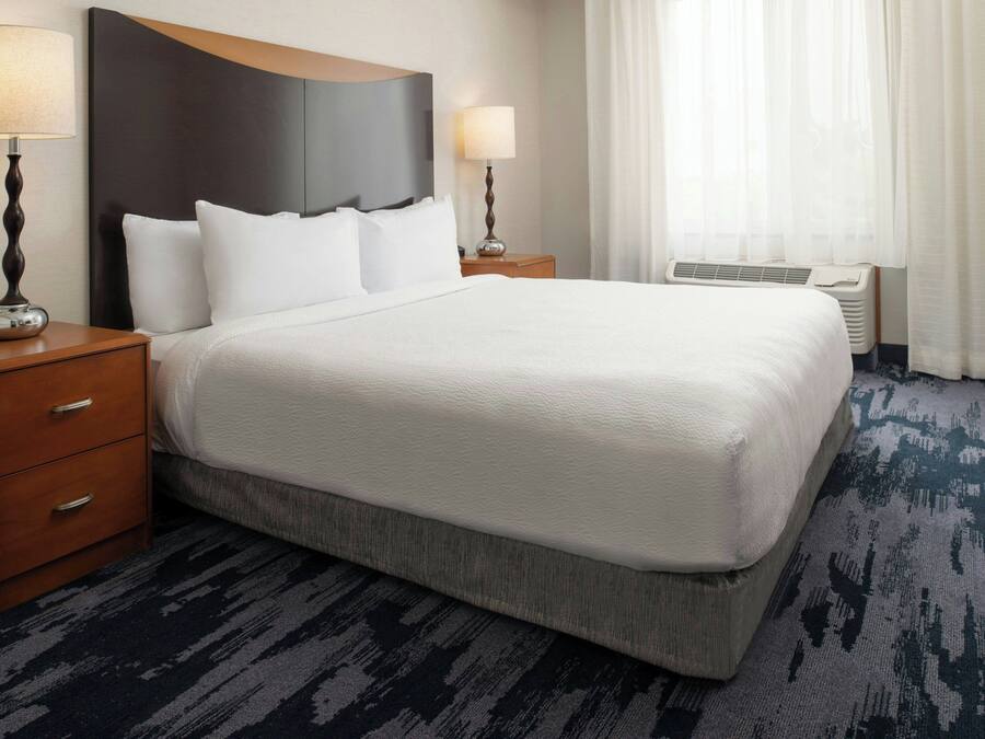 A hotel room with a large bed, two bedside tables with lamps, a wooden headboard, a window with curtains, and a carpeted floor.