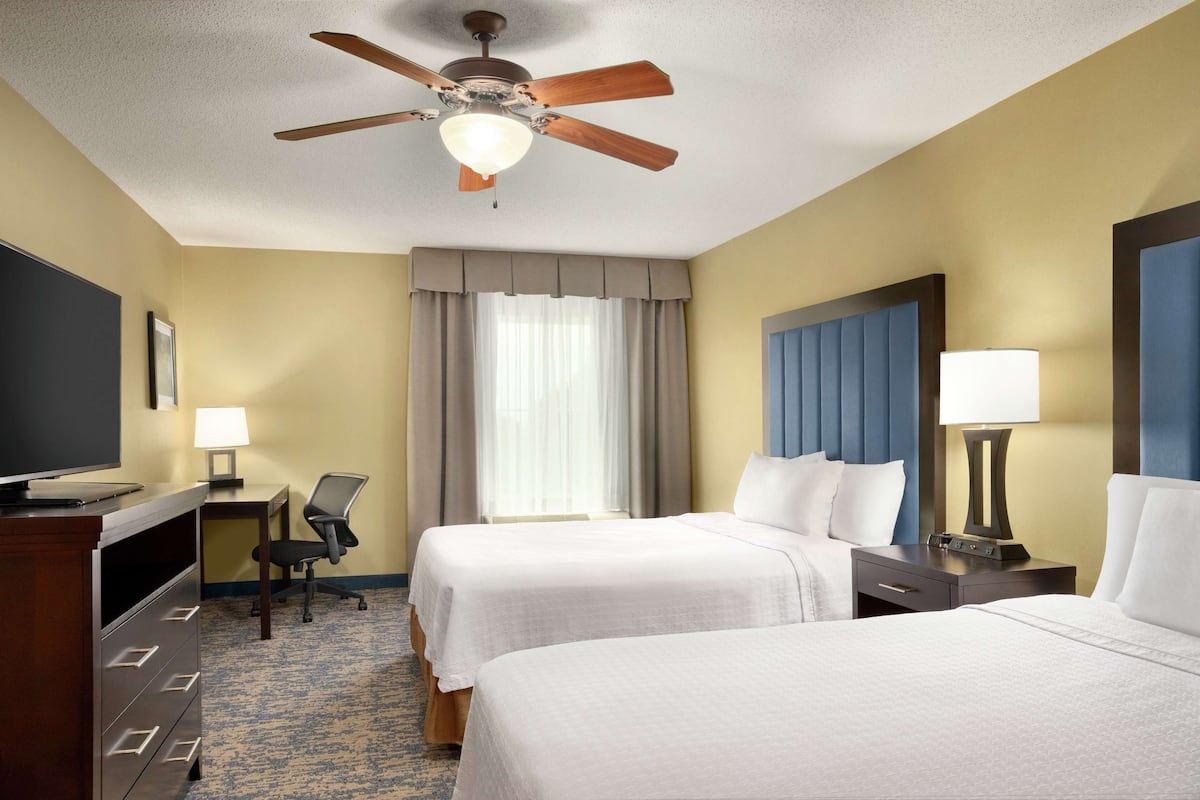Ein Hotelzimmer mit zwei Betten, einem Schreibtisch mit Stuhl, einem Fernseher, einem Deckenventilator und einem Fenster mit Vorhängen.