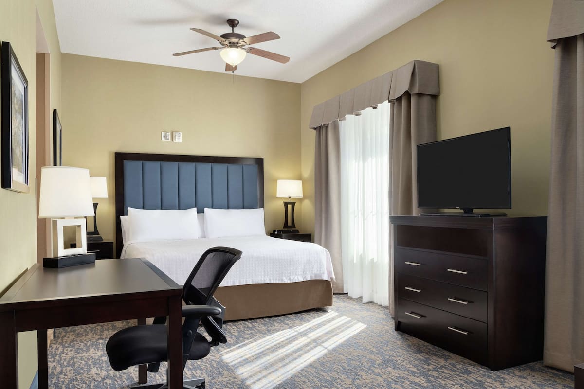 Ein Hotelzimmer mit Bett, Schreibtisch, Sessel, Fernseher, Kommode und Deckenventilator.