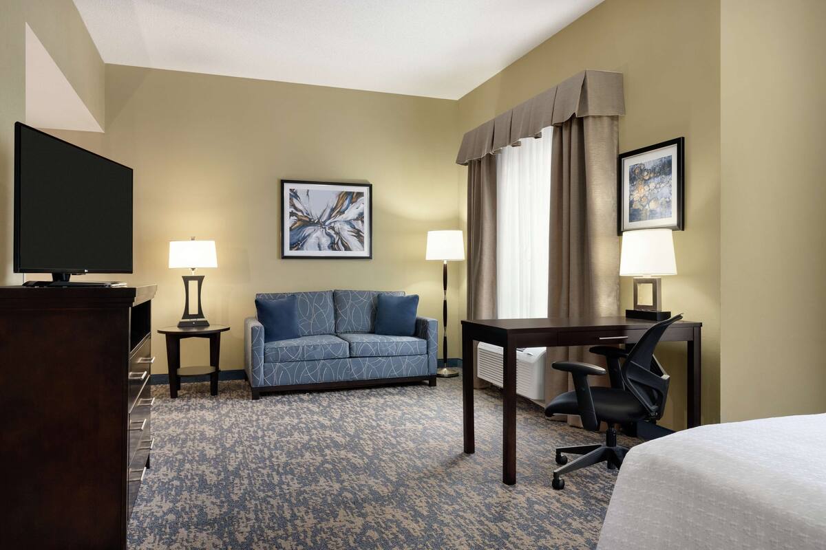 Ein Hotelzimmer mit einem Bett, einem Schreibtisch, einem Sessel, einer Couch, zwei Lampen, einem Fernseher und zwei gerahmten Bildern an der Wand.