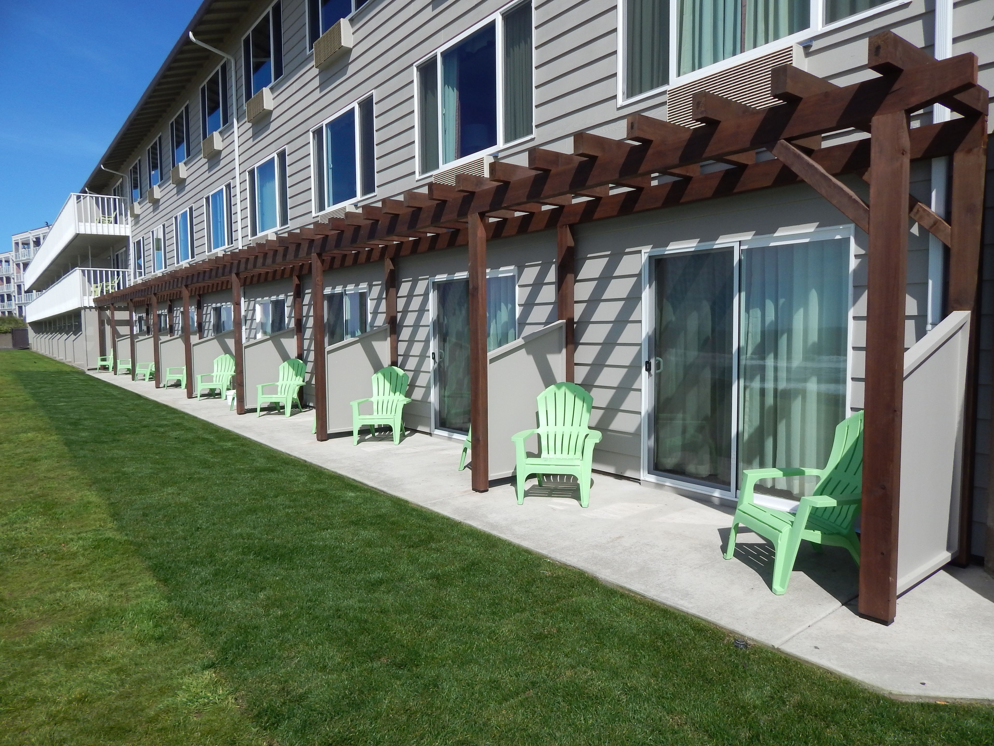 A row of green chairs under a wooden pergola with a glass door, adjacent to a building with multiple windows and a balcony.