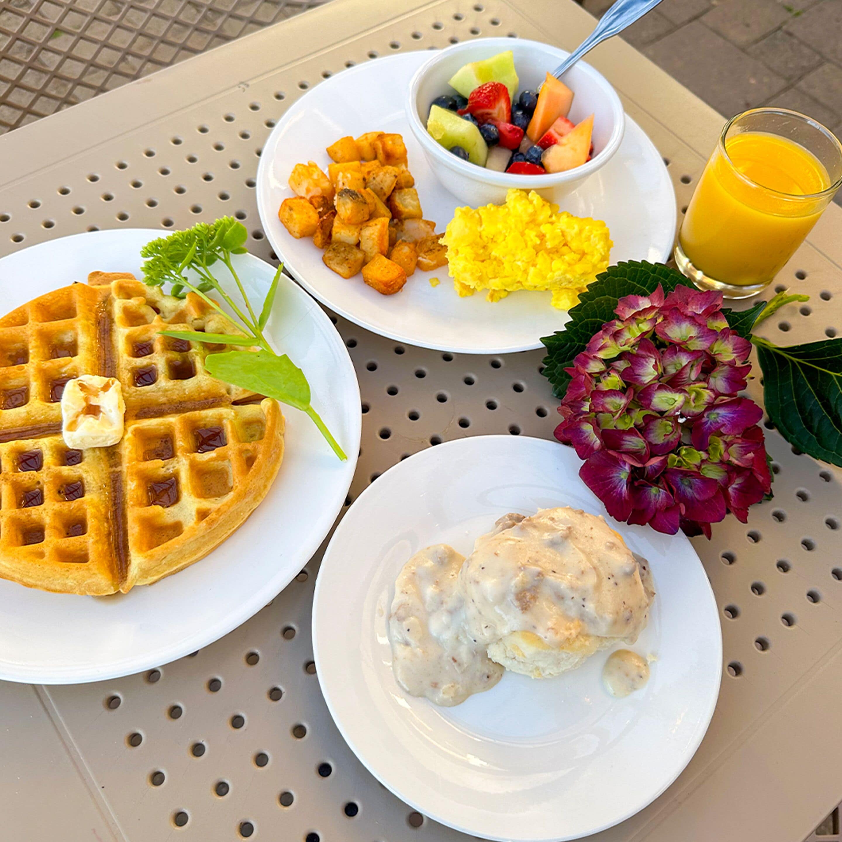 Una mesa con alimentos para el desayuno, como waffles, huevos revueltos, patatas ralladas, ensalada de frutas y un vaso de jugo de naranja.