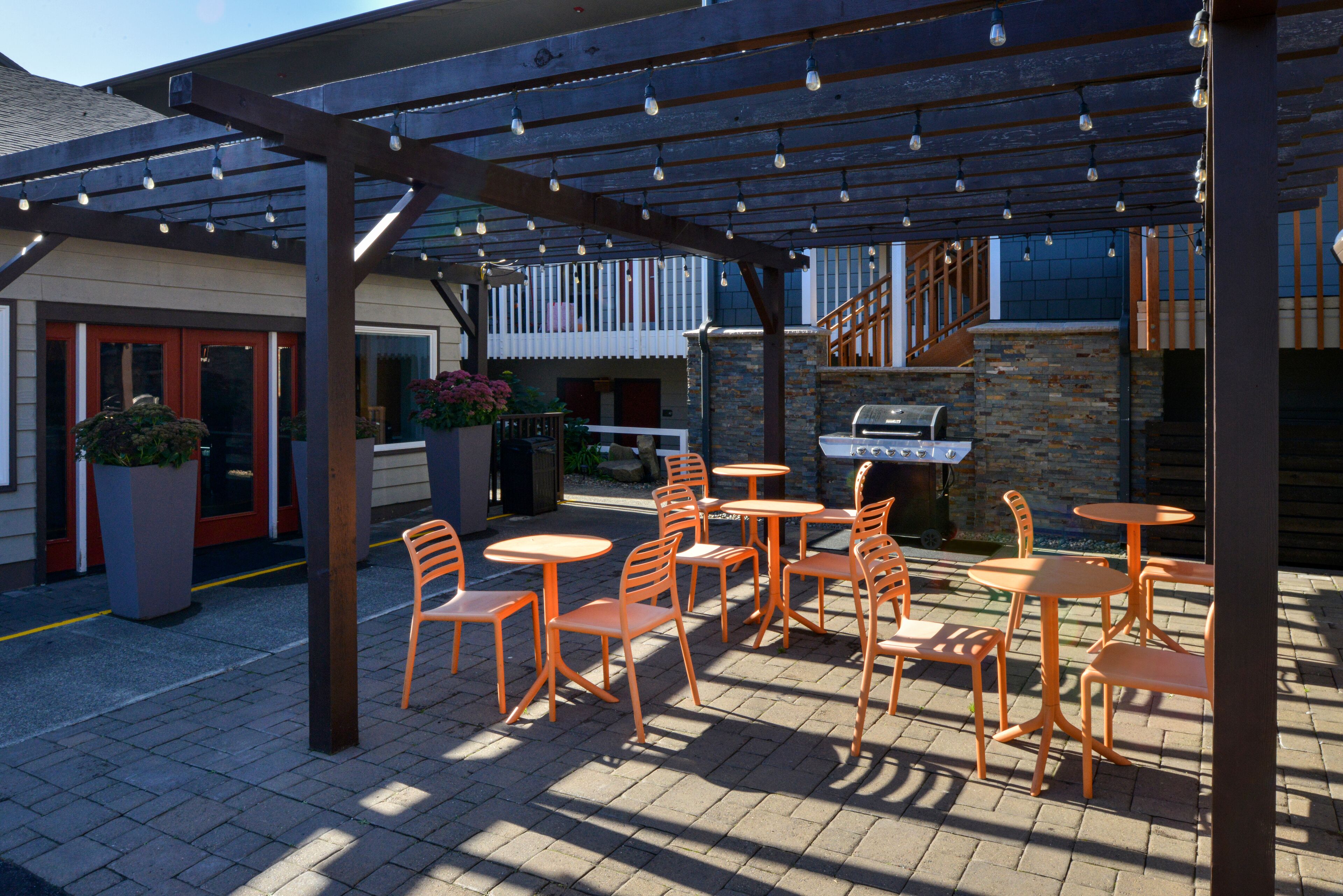 An outdoor patio with orange chairs and tables, a barbecue grill, and a covered area with string lights.