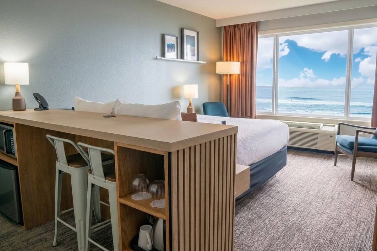 A hotel room with a bed, a desk with two chairs, a view of the ocean, and a window with curtains.