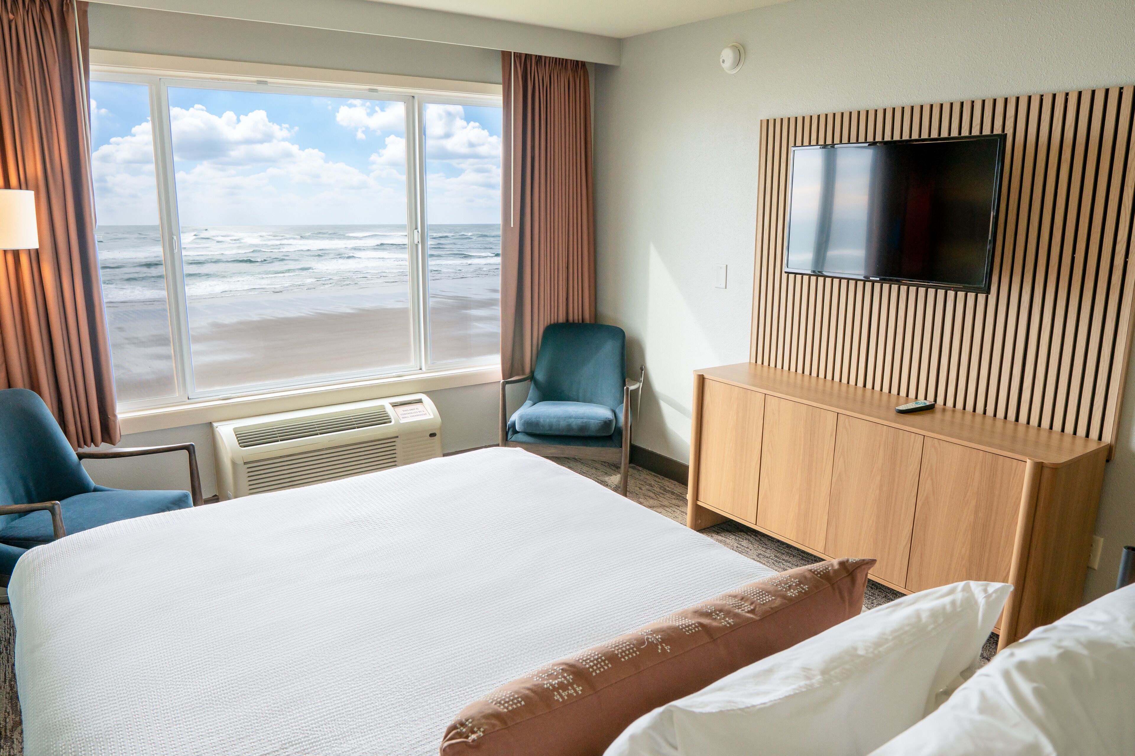 A hotel room with a large bed, a TV mounted on the wall, a chair, and a view of the beach.