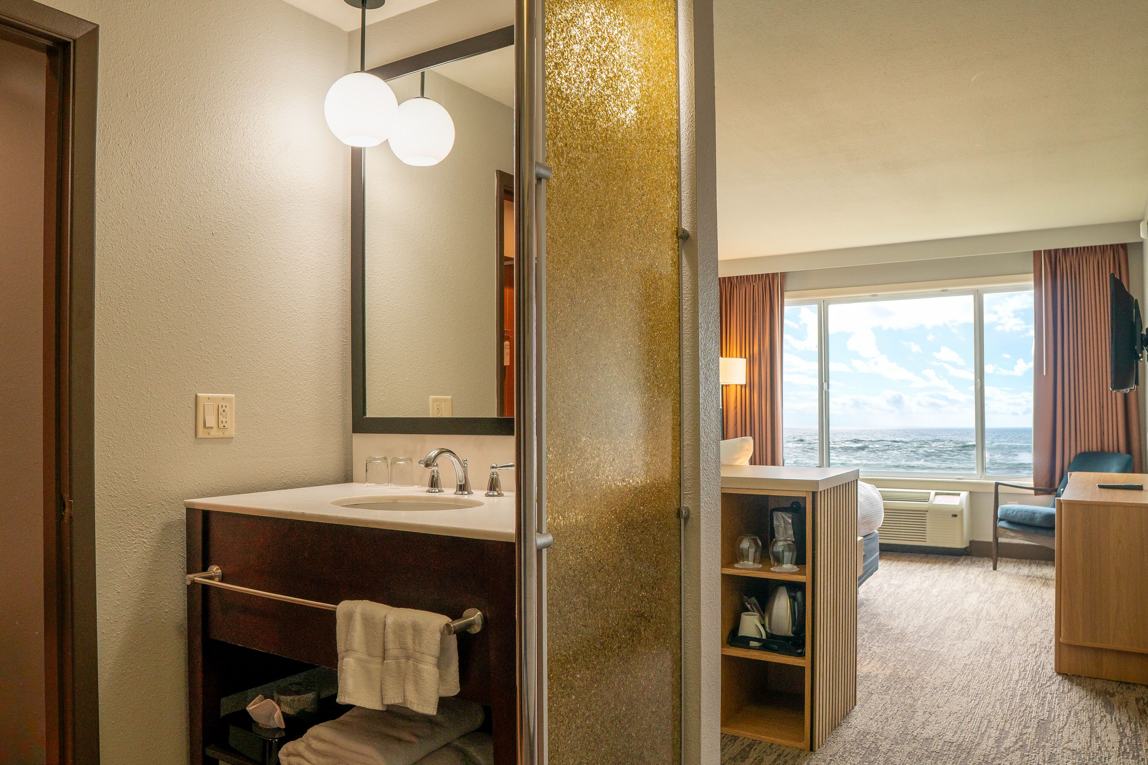 A hotel bathroom with a sink, mirror, towel rack, and a view of the sea through large windows.