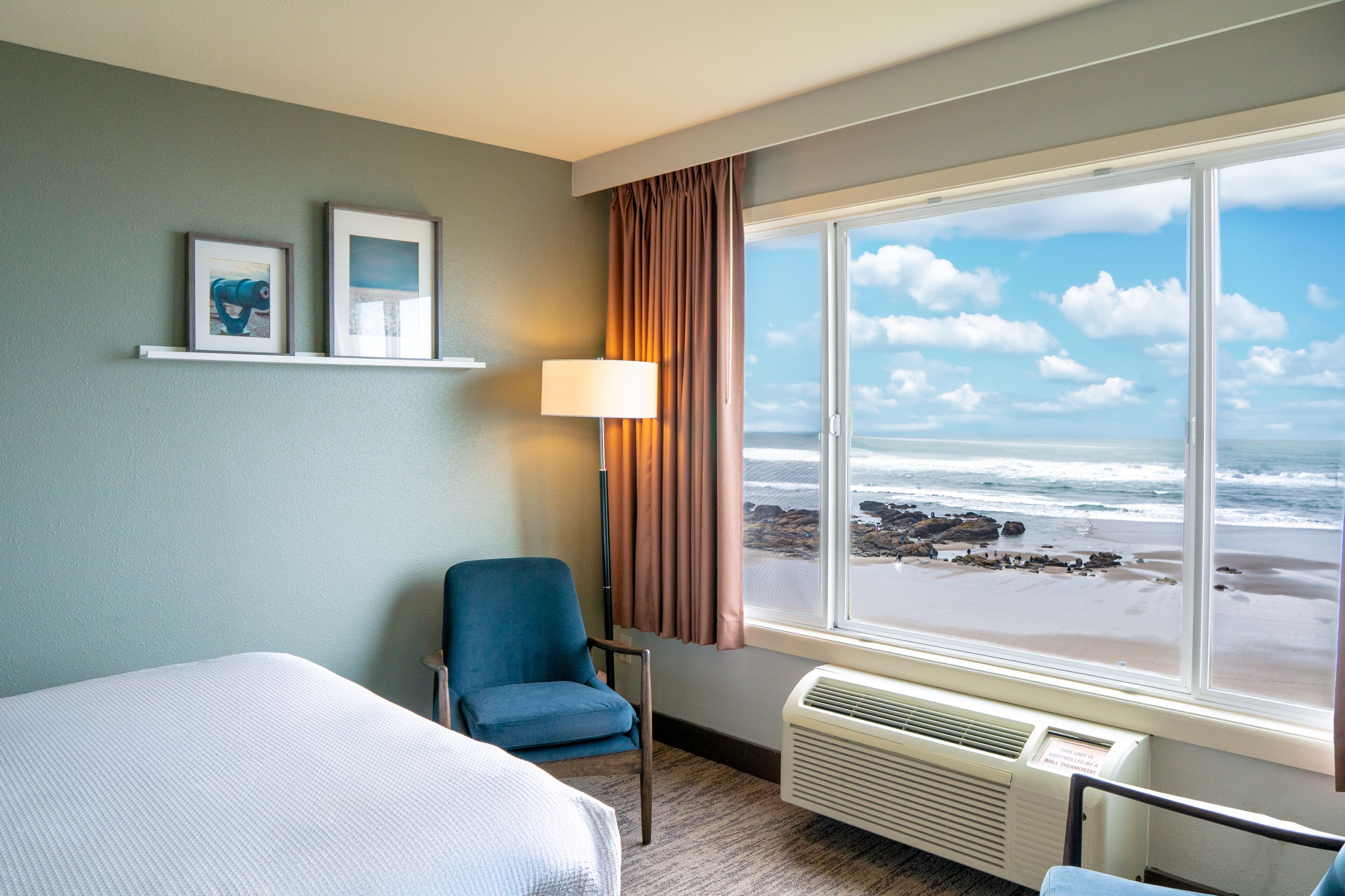 A hotel room with a bed, a chair, a window with a beach view, and two framed pictures on the wall.