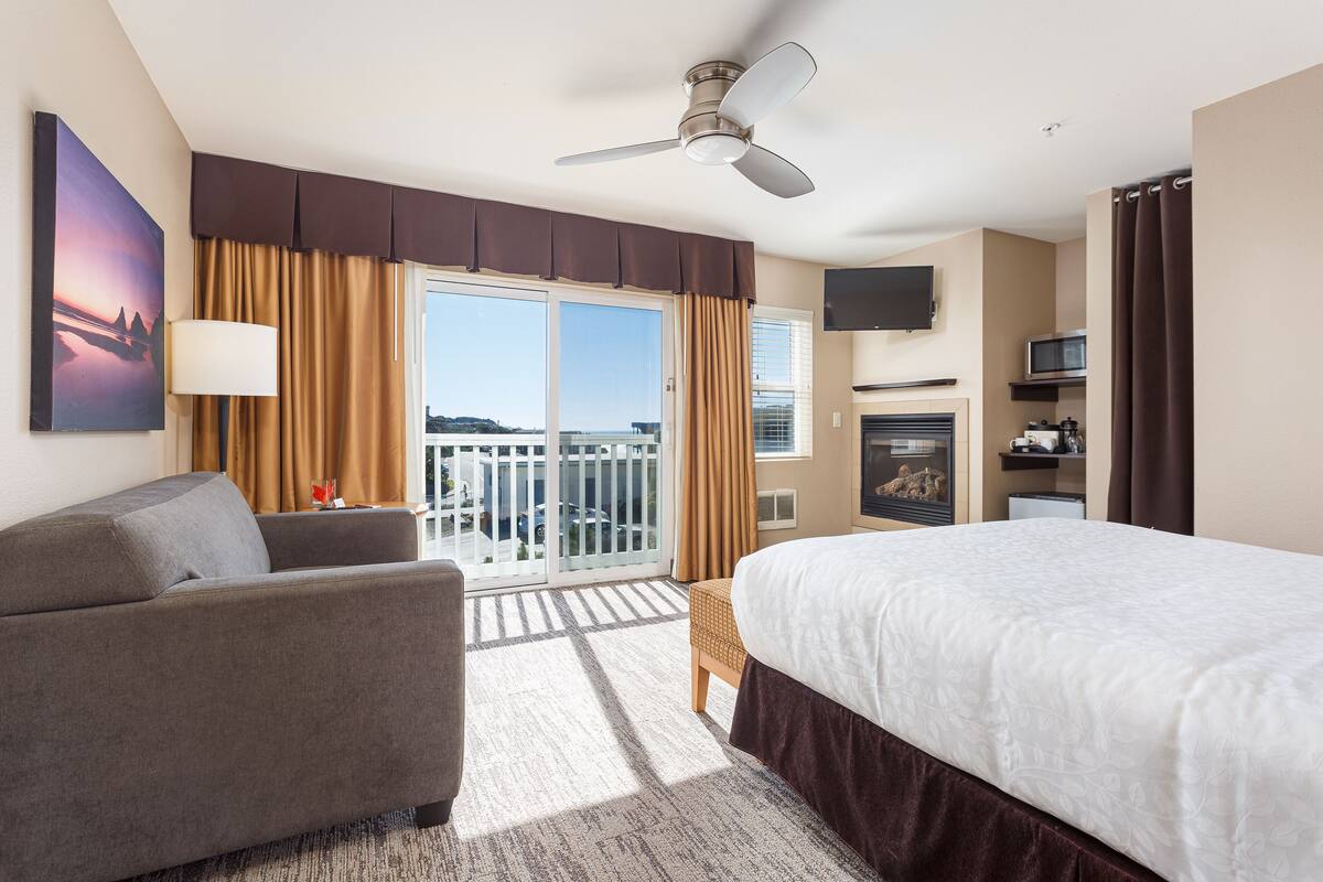 A hotel room with a bed, a sofa, a TV, a ceiling fan, and a balcony with a view.