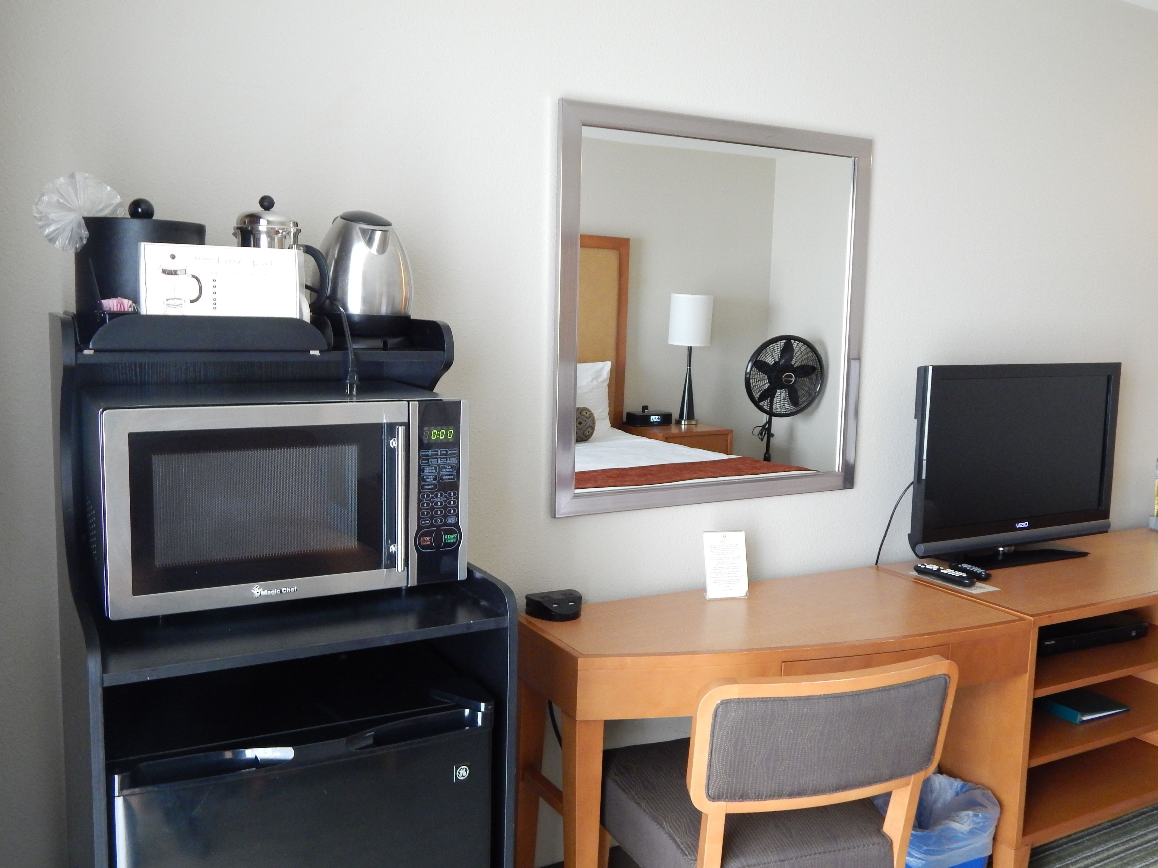 Quarto de hotel com cozinha compacta, incluindo micro-ondas, geladeira e máquina de café. Há uma TV, uma escrivaninha de madeira e uma cadeira.